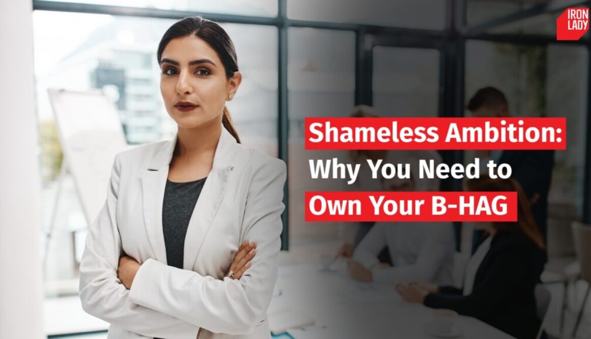 Confident woman in a white blazer standing with arms crossed in an office setting. Text overlay reads: 'Shameless Ambition: Why You Need to Own Your B-HAG.